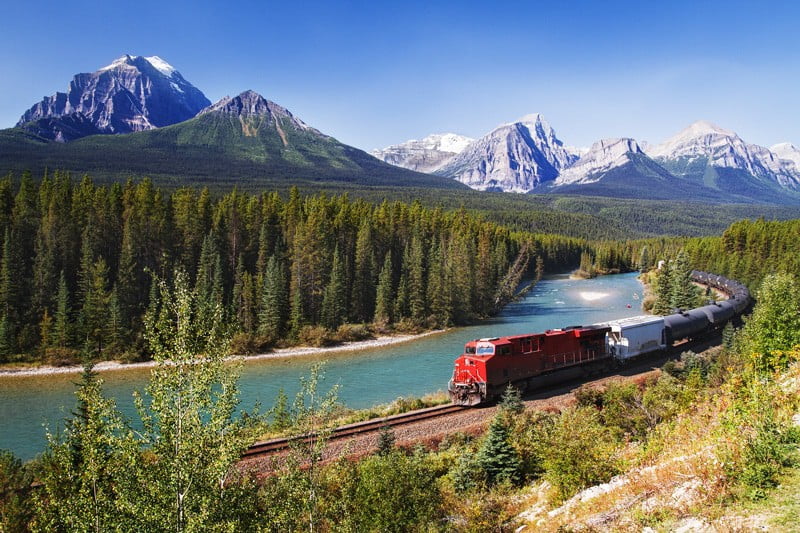 Train with red engine at front going through forest with mountains in the background